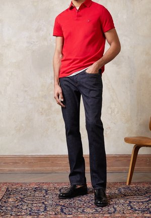 Homme portant un polo rouge, un jean bleu foncé et des chaussures habillées noires, debout sur un tapis à motifs, la main dans la poche, à l'intérieur près d'une chaise en bois.