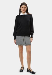 Schwarzer Strickpullover mit strukturiertem Vorderteil, kombiniert mit einem Hahnentrittmuster-Minirock mit Fransenbesatz und Knopfdetails auf der Vorderseite. Schwarze Ballerinas.