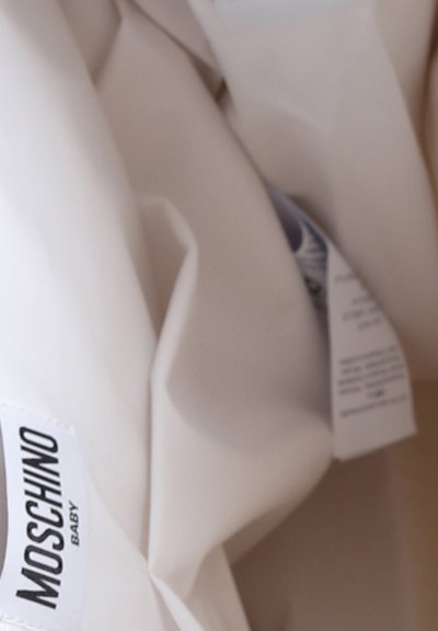 Tessuto bianco con una texture morbida, caratterizzato da un'etichetta nera visibile con la scritta "Moschino Baby". Il materiale appare stropicciato con pieghe sottili.