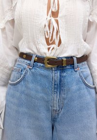 Blouse blanche à lacets rentrée dans un jean en denim bleu clair taille haute avec une ceinture en cuir marron ornée d'une boucle en laiton.