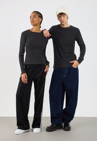 Duas modelos estão lado a lado. Uma veste uma blusa preta de manga longa justa em tecido canelado e calças de treino pretas com riscas brancas. A outra veste uma blusa preta de manga longa e jeans escuros e largos.