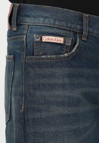 Jeans din denim bleu închis cu un etichetă Calvin Klein pe buzunarul din față, având cusături portocalii și o textură netedă.
