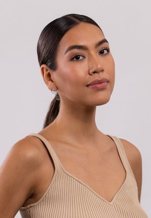 Runde silberne Ohrringe mit einem strukturierten, kreisförmigen Design, vor einem hellen Hautton und einem neutralen, eng anliegenden, gerippten beigen Oberteil.