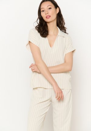 Donna con capelli scuri che indossa un top beige a righe sottili maniche corte e pantaloni abbinati, in piedi davanti a uno sfondo chiaro uniforme.