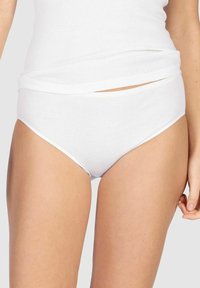 Frau trägt weiße Baumwollunterwäsche und ein passendes weißes Tanktop vor einem schlichten hellen Hintergrund.