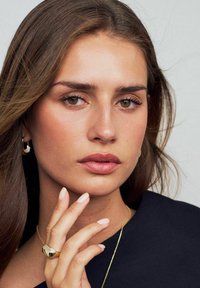 Guldring med texturerad yta på hand, mjuka rosa naglar, enkel guldhalskedja, neutral makeup, brunt hår, bär en mörk topp.