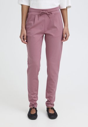 Kobieta ubrana w lawendowe spodnie ze sznurkiem i czarne baleriny, stojąca na tle jednolitego jasnego tła.