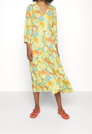 Robe portefeuille florale en vert clair avec de grandes fleurs jaunes et bleues, des manches longues et une jupe à plusieurs niveaux. Portée avec des sandales à brides corail.