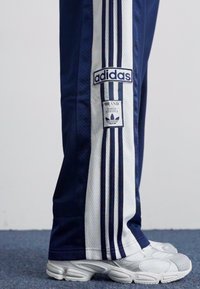 Adidas teplákové nohavice v námorníckej modrej farbe s bielymi sieťovinovými postrannými pruhmi, s označením značky a dizajnom troch pruhov, skombinované so striebornými športovými topánkami.