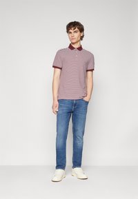 Camisa polo a rayas en granate y blanco, con mangas cortas y cuello. Combinada con jeans azules y zapatillas blancas. Fondo liso.