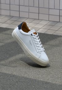 Sneaker en cuir blanc avec un talon bleu marine, bout rond et accents marron contrastés. Dispose de lacets plats et d'une semelle en caoutchouc texturée.
