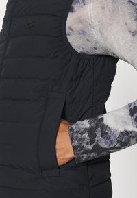 Vesta puffă neagră cu textură matlasată și buzunare laterale. Asortată cu o cămașă cu mâneci lungi, patterned în nuanțe de gri și albastru.