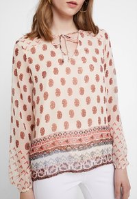 Blusa leggera e trasparente colore pesca con motivo astratto marrone, scollo a V e dettaglio con fiocco, maniche lunghe e bordo inferiore decorato.