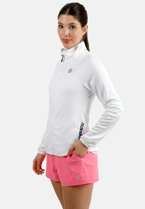 Frau trägt eine weiße Jacke mit Reißverschluss und auffallend pinke Shorts, steht mit einer Hand in der Tasche und ist leicht seitlich gedreht.