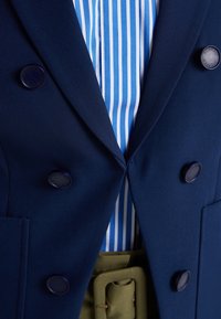 Navy double-breasted blazer with round blue buttons, featuring a light blue and white striped shirt underneath and a green belt accent.