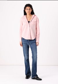 Camisa rosa clara com botões, com colarinho, dois bolsos no peito e mangas compridas, combinada com jeans azuis de perna larga e sapatos pretos.