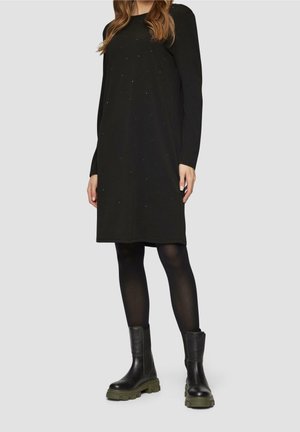 Woman wearing a black long-sleeve knee-length dress with subtle dots, black tights, and black chunky boots with green soles.