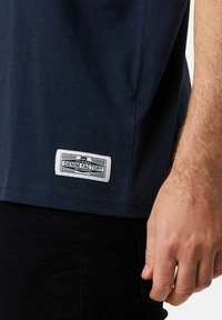 Mörkblå bomullströja med rak nederkant; har en rektangulär vit etikett med texten "DENIM FACTORY" broderad längst ner till vänster.