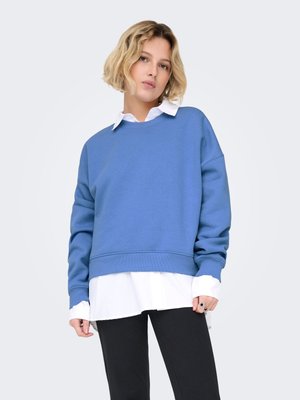 Blue cotton sweatshirt with dropped shoulders, ribbed hem, and a layered white button-down shirt underneath. Paired with black pants.
