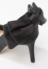 Chaussure noire à talon haut avec finition en satin, ornée d'un grand noeud décoratif sur la bride de cheville, sur fond blanc.
