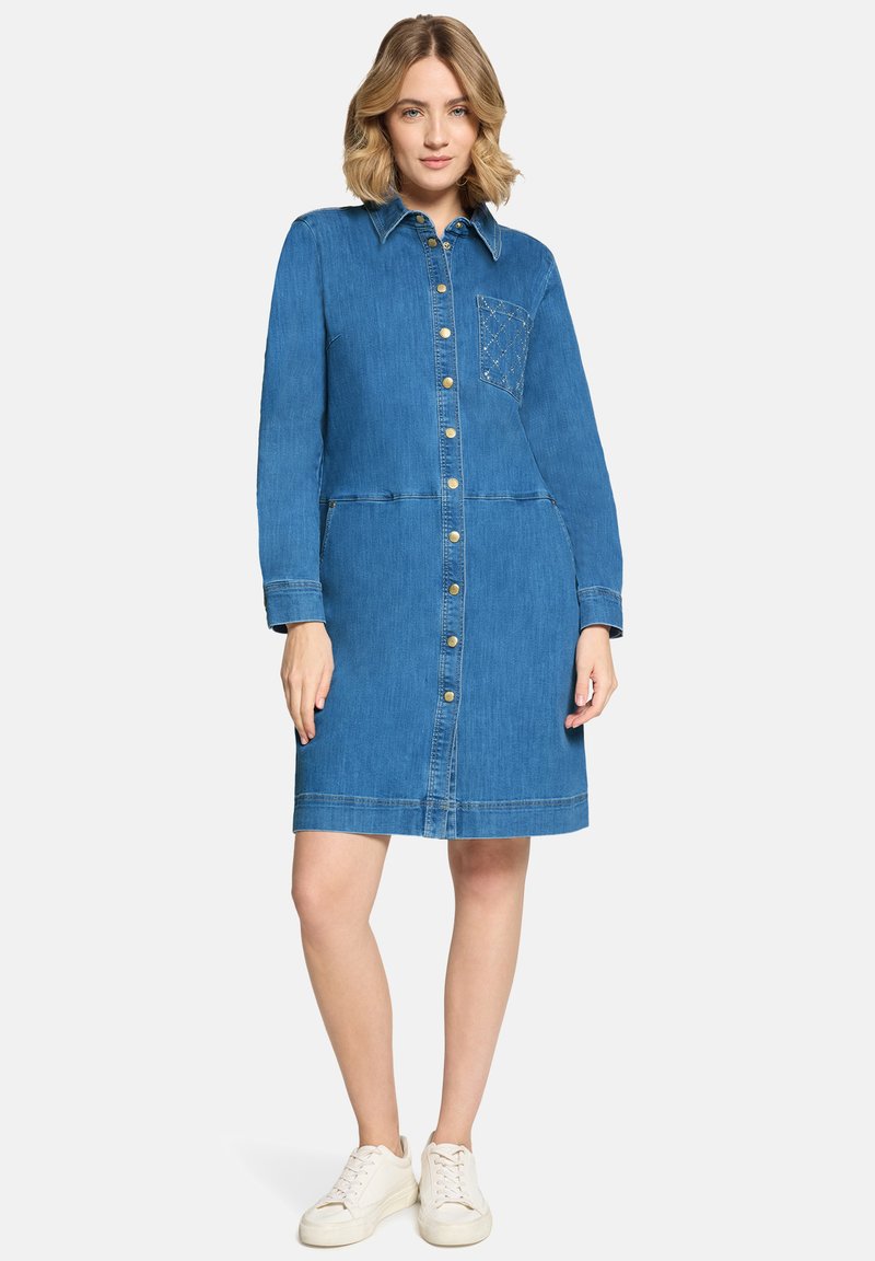 Femme portant une robe en denim boutonnée jusqu'aux genoux, avec manches longues, col, et une poche avant cousue, associée à des baskets blanches.