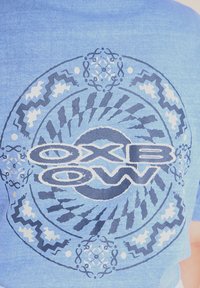 T-shirt in cotone blu con una grafica rotonda caratterizzata da motivi circolari, con il testo "OXBOW" in grassetto bianco e accenti blu scuro.