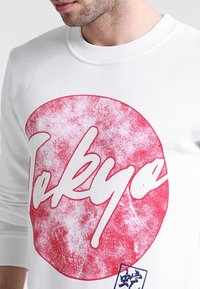 Sweatshirt blanc avec un graphisme circulaire rouge arborant le mot "Tokyo" en lettres blanches majuscules. Tissu lisse avec col et poignets côtelés.