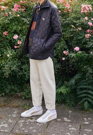 Gepolsterte schwarze Jacke, darunter ein hemd mit tanfarbenem und blauem Muster, cremefarbene Hose, weiße Sneaker mit goldenen Akzenten, vor floralem Hintergrund.