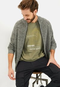 Szary kardigan z teksturą nałożony na oliwkowy t-shirt z żółtym nadrukiem graficznym, w połączeniu z ciemnymi spodniami cargo. Osoba siedząca na stołku.