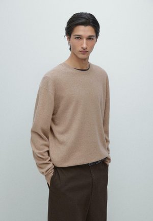 Maglione lavorato a maglia color marrone chiaro con collo rotondo e maniche lunghe, indossato sopra uno strato di base scuro, abbinato a pantaloni marrone scuro.