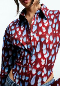 Cămașă cu nasturi, cu model roșu și albastru, cu guler, având un design mulat și motive distincte asemănătoare cu pene pe un material texturat.