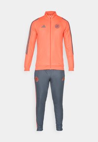 Orangenes Sportjackett mit grauen Akzenten und Streifen, kombiniert mit grauen Hosen, die orangefarbene Details und Logos aufweisen. Mit Reißverschluss, sportlicher Schnitt.