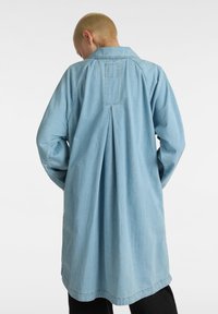 Camicia in denim azzurro chiaro, lunga, con colletto, dettaglio di piega sul retro e maniche lunghe. Il tessuto ha una texture liscia ed è leggermente over-size.