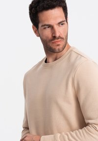 Sudadera de cuello redondo en color beige claro, fabricada con un tejido suave. Presenta un cuello y puños de canalé, con una textura suave y un diseño minimalista.