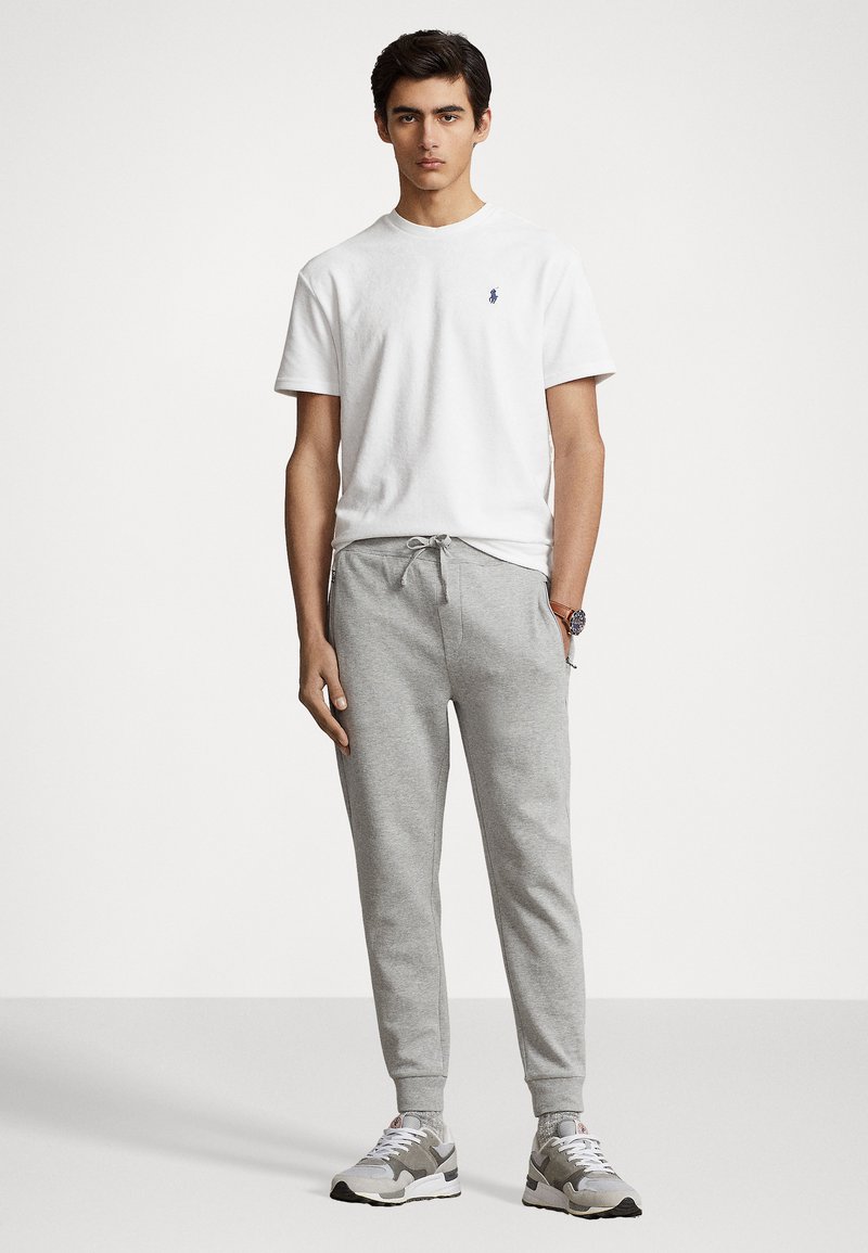 Witte katoenen t-shirt met een klein logo, gecombineerd met lichtgrijze sweatpants. Het model draagt grijze en witte sneakers, wat een casual uitstraling geeft.