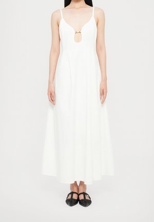 Woman wearing a sleeveless white maxi dress with thin straps, a gold clasp detail at the deep V neckline, and black pointed-toe shoes.