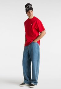 Camiseta roja de manga corta, corte relajado, con logo en el pecho. Combinada con vaqueros de pierna ancha de color azul claro y un gorro negro con diseño gráfico blanco.