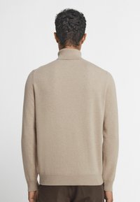 Beige, stickad tröja med hög krage, ribbade ärmslut och fåll. Slät textur med en avslappnad passform, visad bakifrån.