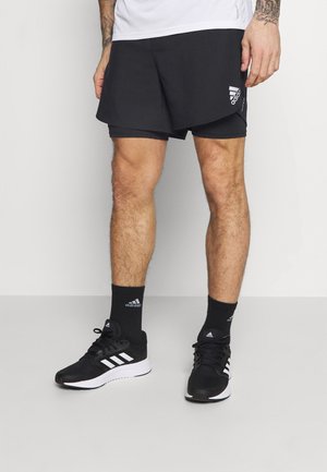 Mustat urheilushortsit kerroksellisella muotoilulla, yhdistettyinä mustiin tennareihin ja sukkiin, joissa on valkoiset Adidas-raidoitukset ja logokorostuksia.