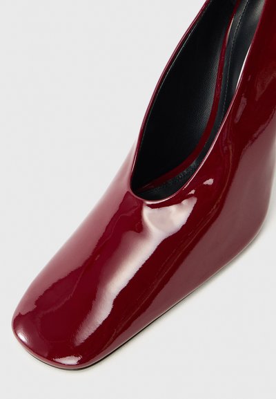Pantofii cu toc din piele lac burgundă, cu vârf pătrat, siluetă elegantă și textură netedă. Prezintă o decupare adâncă pe lateral pentru vizibilitatea piciorului.
