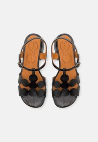 Sandali in pelle nera con accenti decorativi circolari, cinturino regolabile e soletta beige. Superficie texturizzata con design aperto sulla punta.