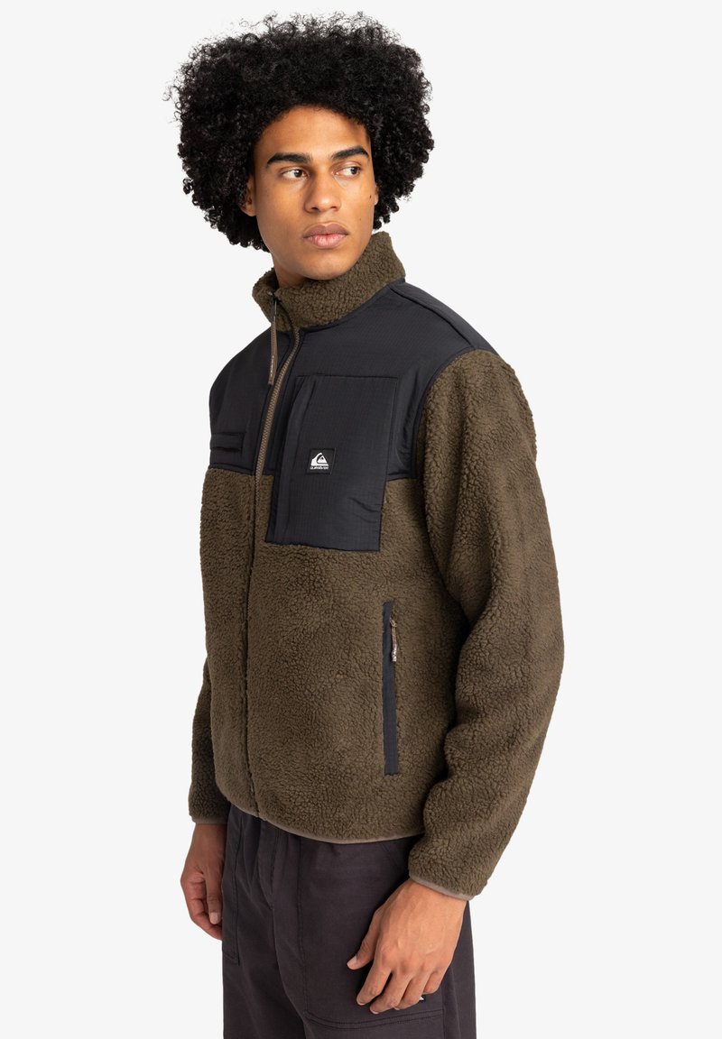 Fleece jacket in brown with black nylon accents. High collar, front zip, two zippered pockets, and two chest pockets with flaps.