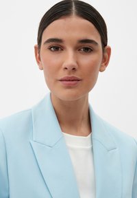 Blazer azzurro chiaro con rever a taglio, abbinato a una maglietta bianca. Il tessuto liscio e il design strutturato mettono in risalto linee pulite e taglio moderno.