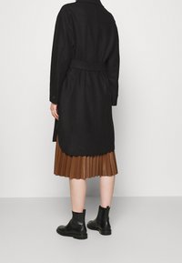 Cappotto di lana nero con vita cinturata, caratterizzato da un collo alto e spacchi laterali, abbinato a una gonna marrone plissettata e stivali neri alla caviglia.