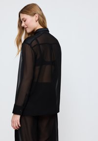 Donna con capelli lunghi che indossa una camicia nera trasparente a maniche lunghe sopra biancheria intima nera visibile, in piedi contro uno sfondo chiaro e semplice.