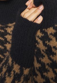 Hand met natuurlijke nagels, gedeeltelijk bedekt door een zwarte, donzige gebreide mouw over een bruin-zwart hondentandpatroon trui.