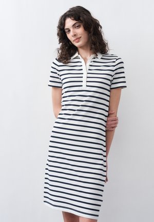 Γυναίκα με σγουρά μαλλιά που φοράει λευκό φόρεμα μέχρι το γόνατο με μαύρες οριζόντιες ρίγες και γιακά με κουμπιά, στέκεται μπροστά σε απλό φόντο.