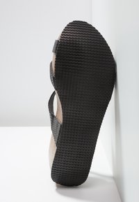 Sandale avec semelle en caoutchouc noire texturée et brides en tissu gris, positionnée debout contre un mur blanc sur une surface blanche.