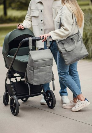 Šedý látkový batoh připevněný k černému kočárku s tmavě zelenou korbou. Osoba drží odpovídající šedou tašku, oba z textilního materiálu.