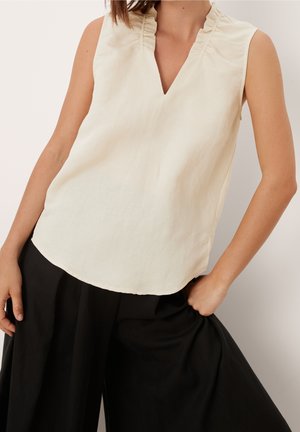 Kremowa bluzka bez rękawów z falbanką przy kołnierzu, noszona z luźnymi czarnymi spodniami, ręce opierające się na biodrach, na jednolitym jasnym tle.